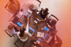 An aerial view image of eight people seated around a desk. Each person seems to have either a laptop, a phone or a tablet in front of them. A pink and orange gradient is overlaid on the image.