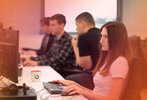 A photo of Ocuco employees sitting at their desk. At the front of the image is a woman in a white t-shirt. A pink gradient is overlaid on the image.