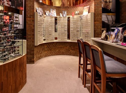 Image of Davis Eyecare’s practice with glasses on a semi-circular back wall. A row of three elegant chairs faces into a countertop of the right-hand side of the image, and merchandising showcasing different frames branding throughout the image. 