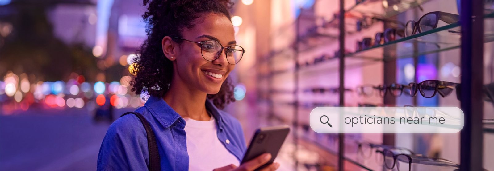 Smiling woman wearing eyeglasses stands in front of an optical store display while using her phone to search for "opticians near me".