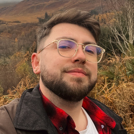 Shane Quilty is a young man with short brown hair and a neatly groomed beard takes a selfie outdoors. He wears gold-rimmed glasses, a small hoop earring, and a nose ring. He is dressed in a red plaid shirt layered under a dark jacket. Behind him is a scenic autumnal landscape with hills, trees in fall colors, and overcast skies.