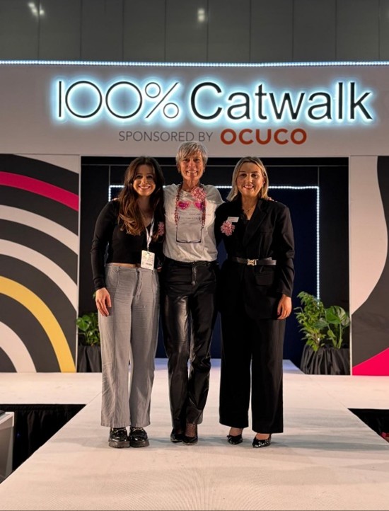 Three Optical Woman Association (OWA) members, including Danielle Martin, standing together on the catwalk stage at 100% Optical 2025. Behind them, the sign reads “100% Catwalk Sponsored by Ocuco” with colorful curved patterns on the backdrop.