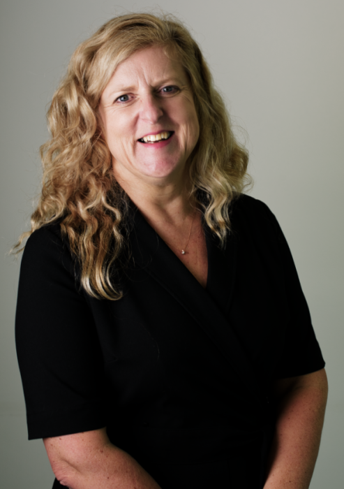 Portrait of Dr Alicia Thompson, smiling woman with long wavy blonde hair wearing a black dress, photographed against a neutral grey background.