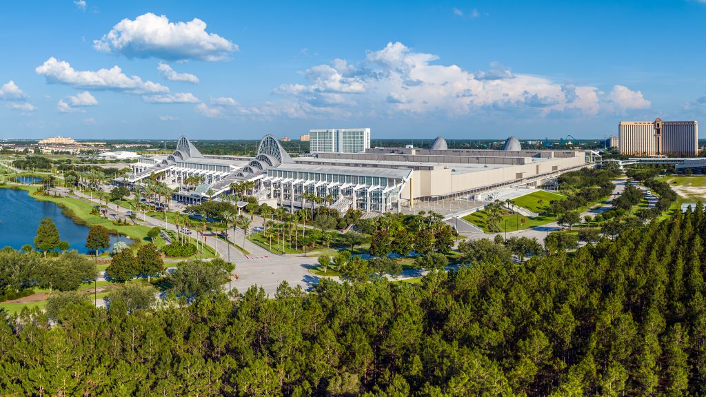 Aerial view of the Orange County Convention Center in Orlando, Florida, surrounded by palm trees, landscaped walkways, and a nearby lake under a bright blue sky.